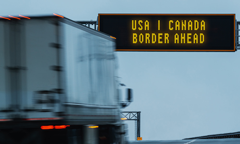 A semi truck drives toward a sign indicating the USA/Canada border