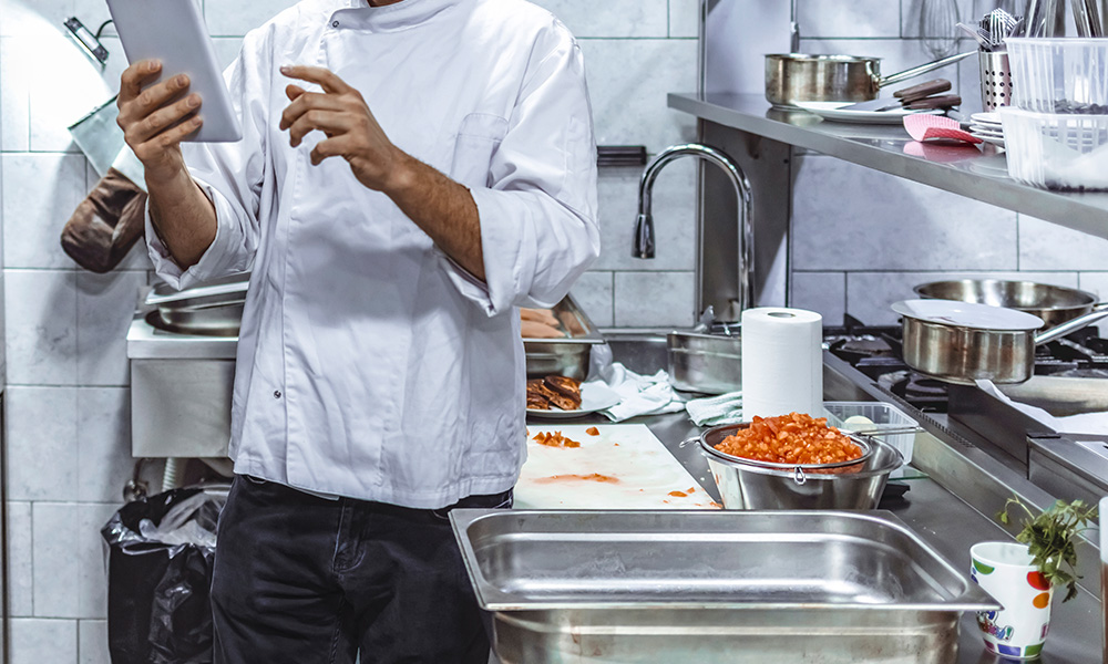 A chef stands in a kitchen holding a tablet