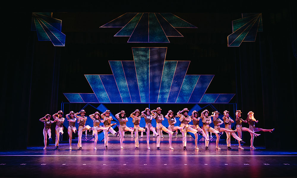 a stage where 18 dancers are all in a row, wearing identical gold pants, vests and shirts, holding top hats as they all dance in a production A Chorus Line