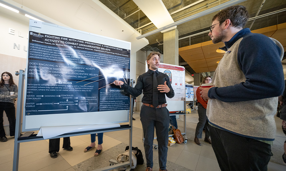 A student points to a poster presentation to explain their work to an onlooker.