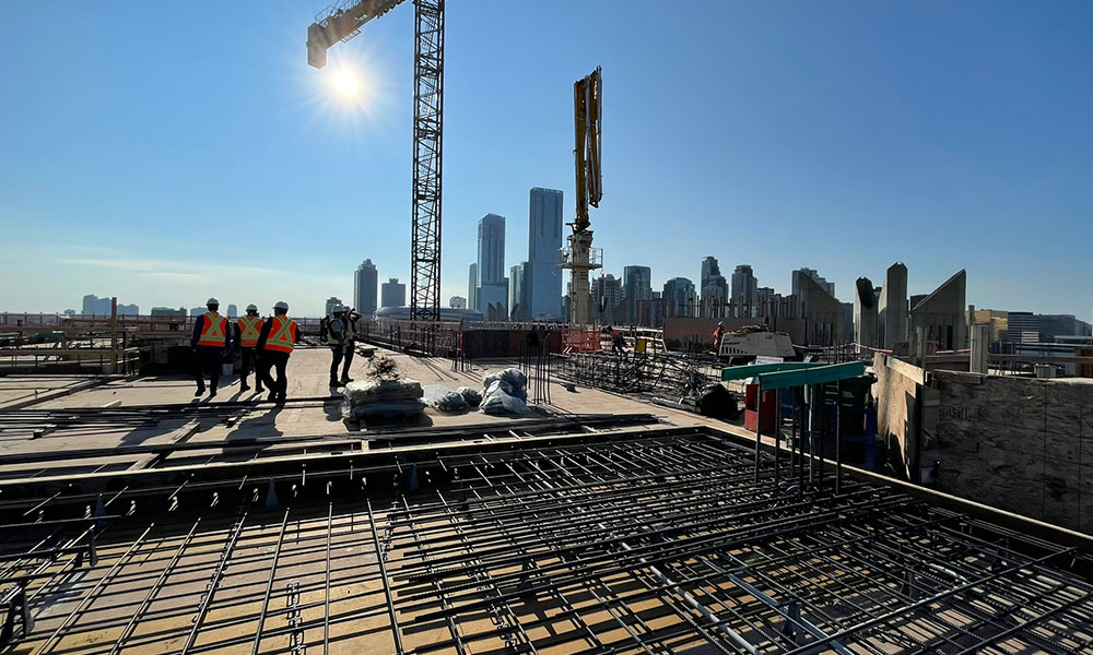 On the top of the building, which is concrete and rebar, people stand on the roof in safety gear and there's a crane in the frame