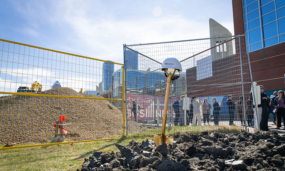A close up of a shovel in a pile of dirt, the handle is upright and a hardhat balances on top of it.