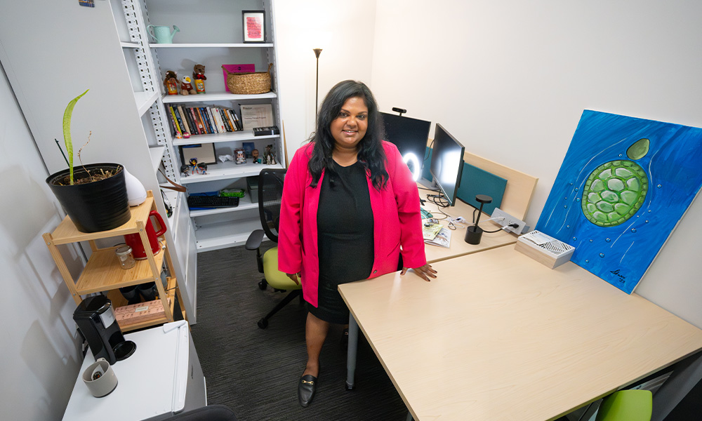 Soni Dasmohapatra stands behind her desk, wearing a black dress and a pink blazer.