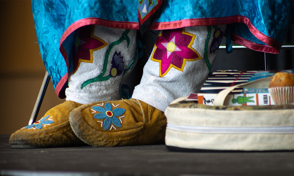 A photo of feet wearing mocassins underneath the edge of a ribbon skirt, alongside a book bag and a muffin