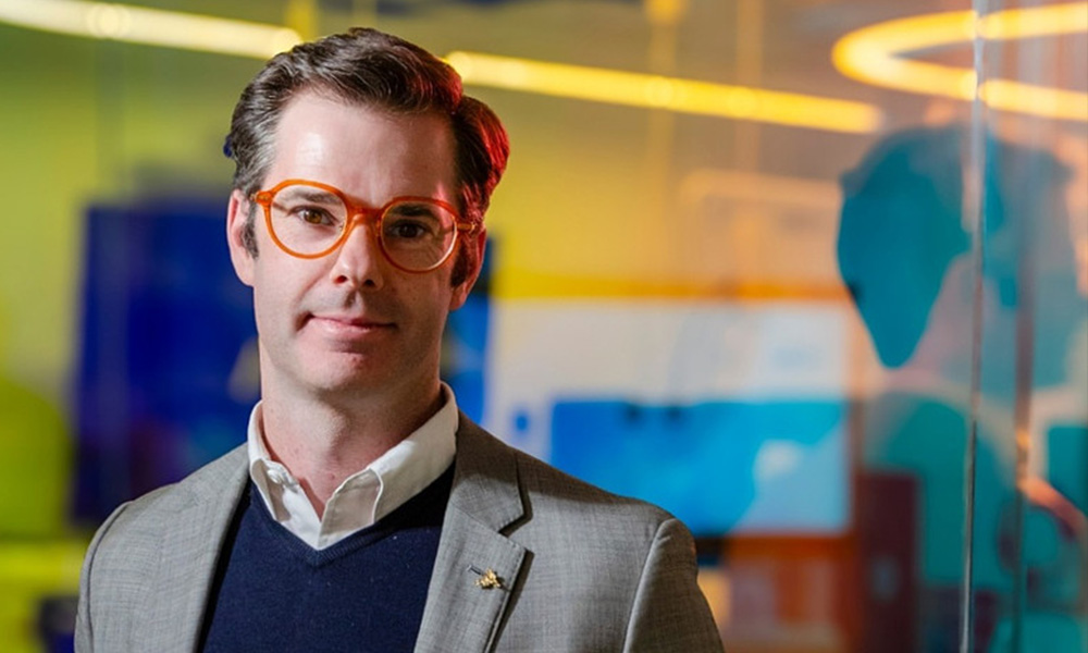 Ryan Payne stands in front of a glass window with neon lighting behind him