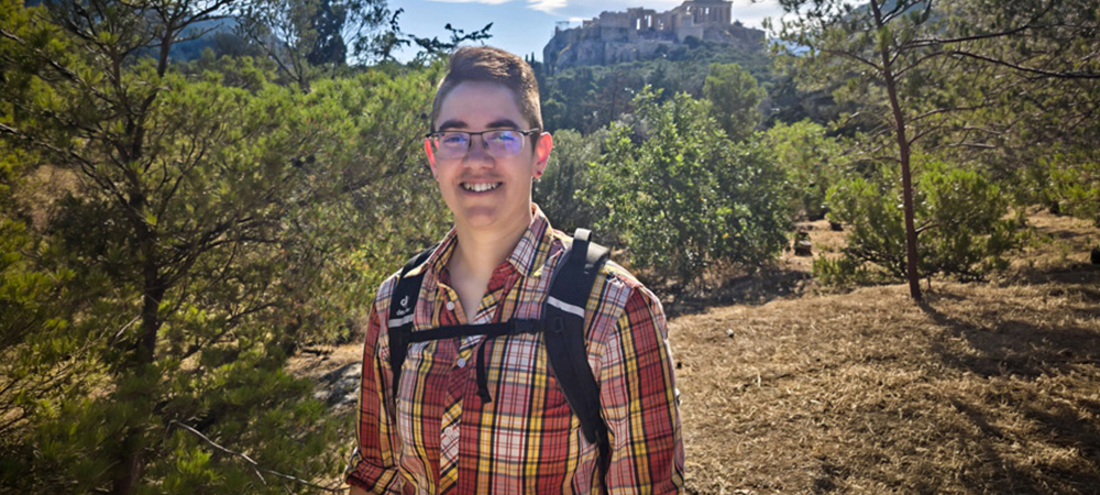 Kat Furtado stands outdoors on a hill with trees and ruins in the background