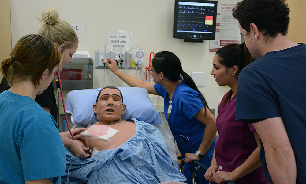 Five students stand together around a hospital bed where a computerized mannequin is laying, hooked up to a monitor and wearing oxygen.