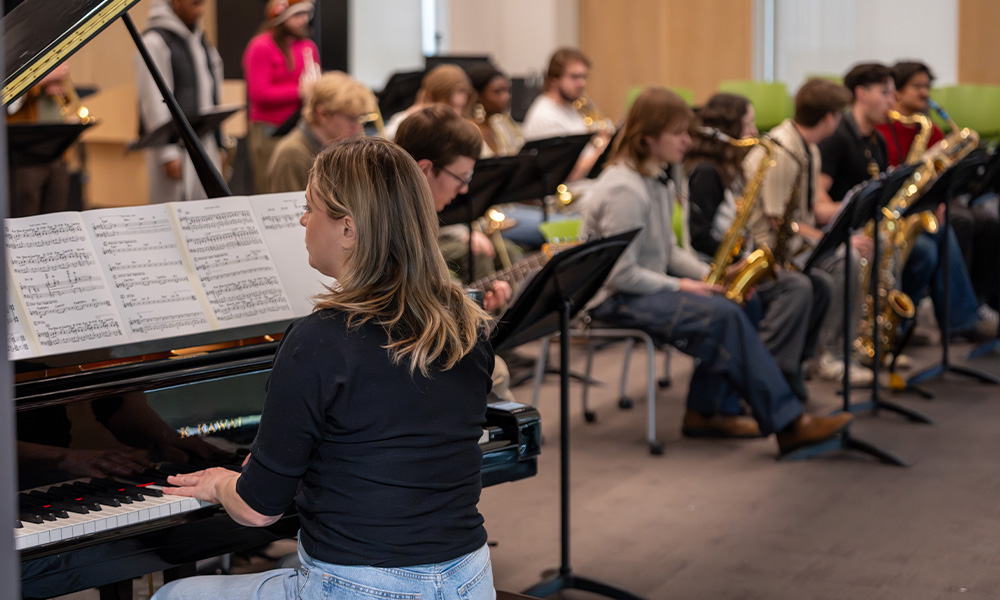 A woman plays a grand piano, and students with various instruments are in the background.