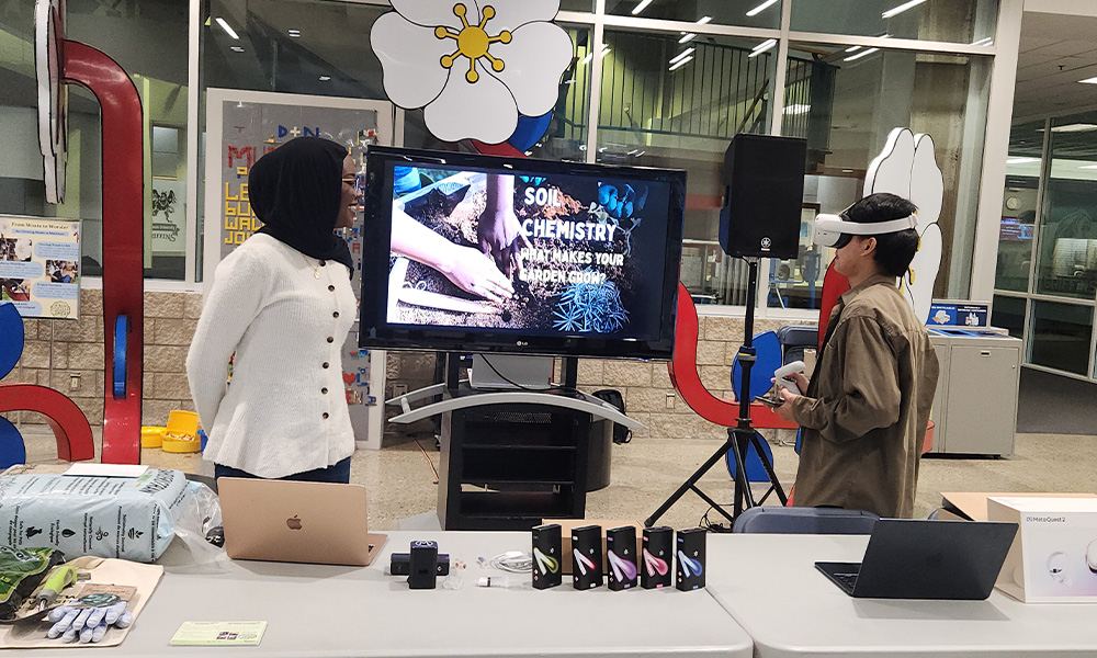 Two students stand at a folding table, one wearing VR goggles.