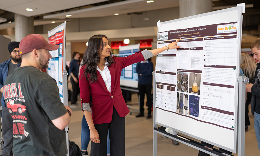 A student in a red blazer gestures to a poster presentation in explanation to an onlooker.