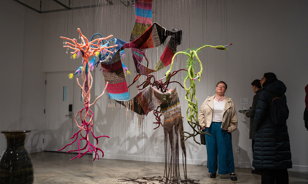 People stand around an intricate, colourful mix of fabrics and textiles hanging from the ceiling of the MAG on campus.
