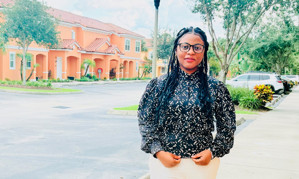 A woman wears a black patterned shirt and white pants and stands in front of a parking lot, a condo building behind her.