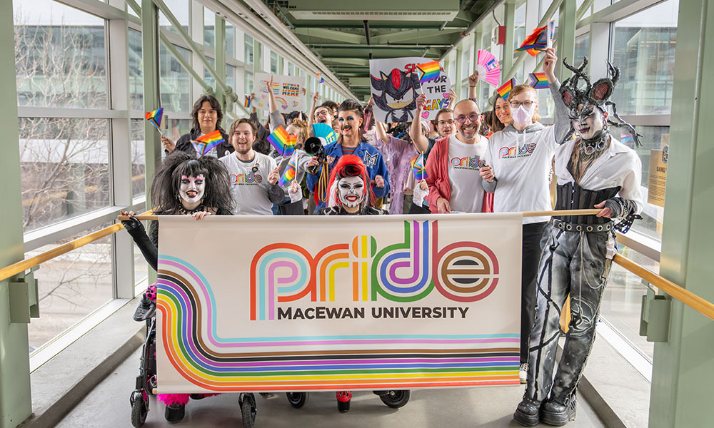 A large crowd marches across the 109 St. pedway, with a Pride banner being held up by the people in front.
