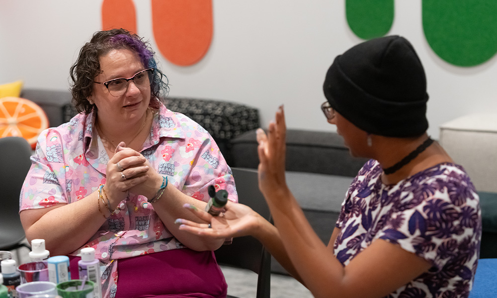 Jessica Scalzo sits with a student, listening intently as the student speaks, both sitting at a table covered in paints and crafting materials.