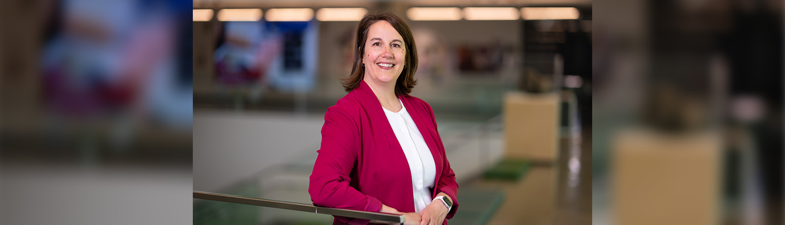miling person in a magenta blazer and white top, leaning on a railing in a modern office with a blurred background