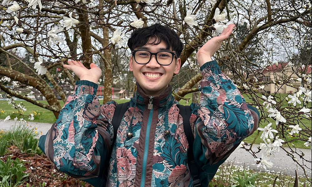 A man stands in front of a blossoming tree holding his hands up on either side of his face