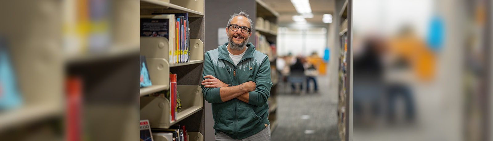 A man in glasses leans against a metal bookstack smiling and with his arms crossed