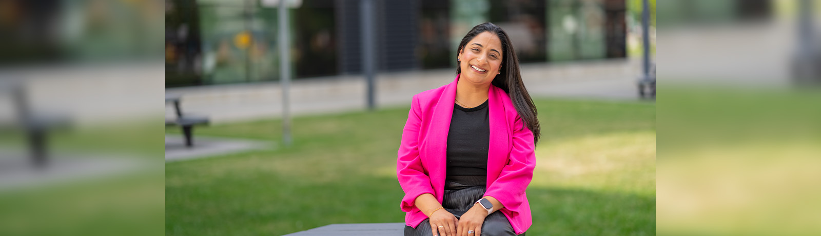 Nooshin Walji wears a hot pink blazer over a black dress, and sits on a picnic table outside of Building 9, smiling.