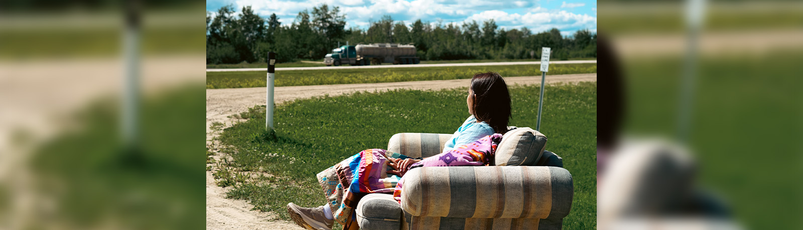 Etienna Moostoos-Lafferty wears a ribbon skirt and sits on a couch along the side of the highway, looking away from the camera.