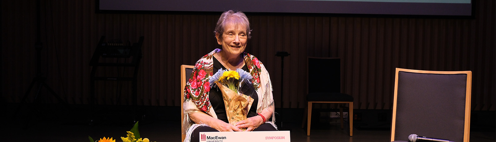 Myrna Kostash sits on stage holding a bouquet of flowers