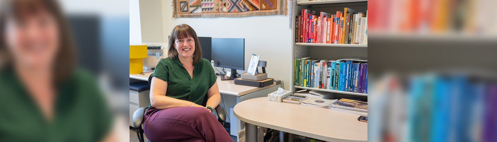 A woman sitting in a chair in an office, in front of a computer on a desk and a bookcase full of books organized by colour on one side of the room.