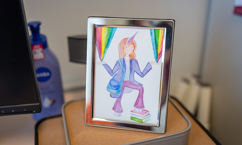 A watercolour painting of a woman with a unicorn horn coming out of her forehead. She is standing on a stack of books, with hooved feet, and rainbows are coming out of her hands.