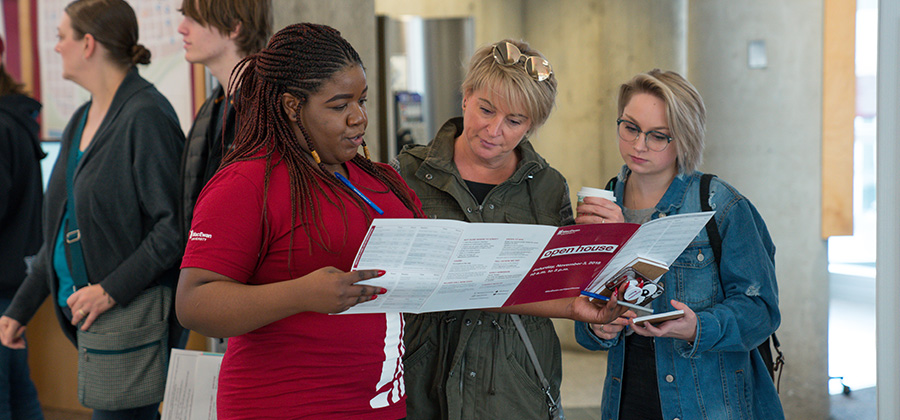 Three people stand together looking at a brochure being held by one woman who is wearing a maroon t-shirt with a MacEwan logo on it