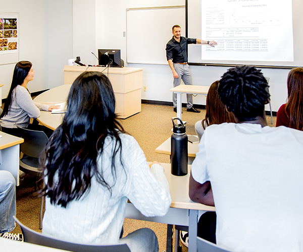 students sitting in classroom with teacher at the front pointing to whiteboard