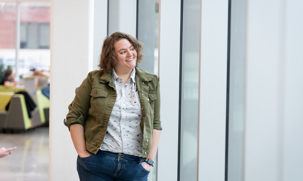 Andi leaning against a window smiling, hands in pockets of jeans