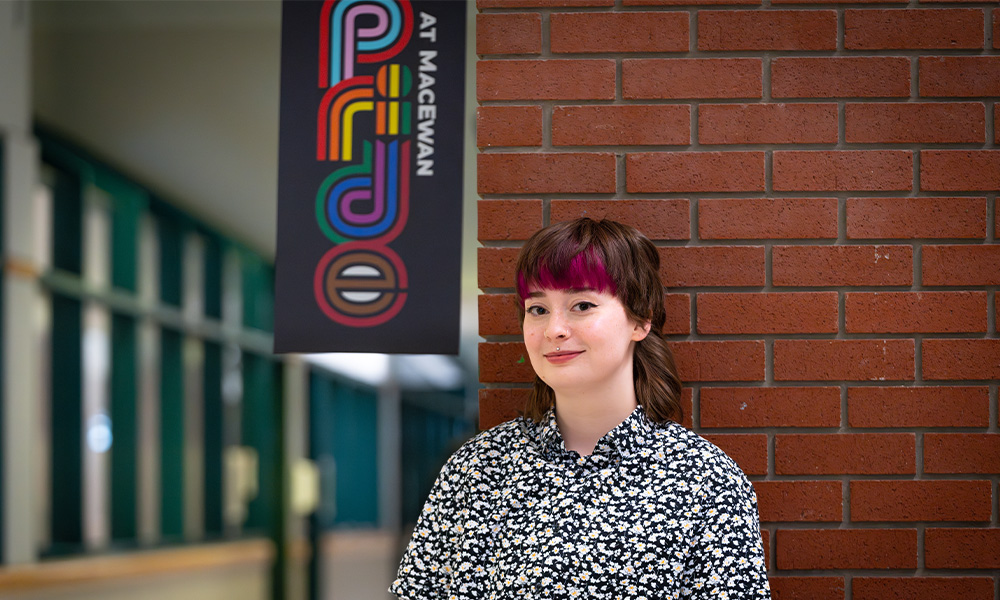 Alana standing in front of a brick wall, a Pride At MacEwan banner is above her head.