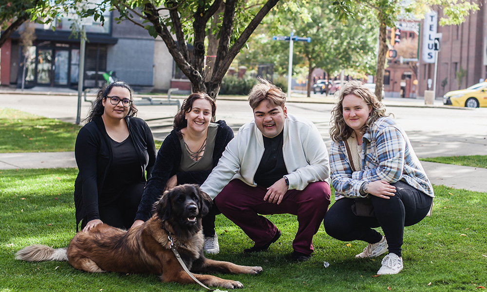 Four people crouch on the grass with a large brown and black dog, all smiling.