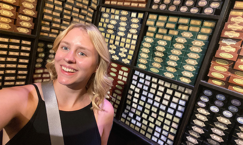 Brynne, a young woman with blonde hair and blue eyes, smiles at the camera in front of a wall displaying many small boxes of items.