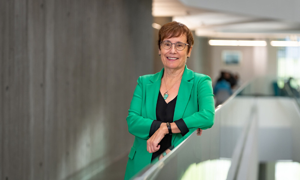 Dr. Annette Trimbee wears a green blazer and leans against a glass railing in Allard Hall.