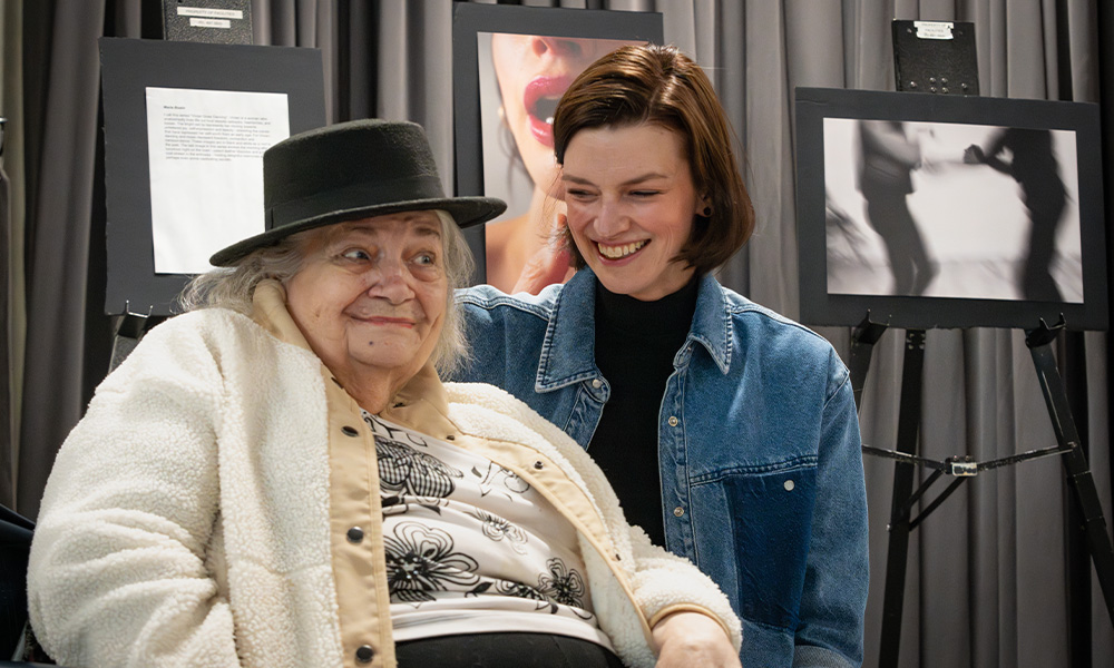A senior woman wearing a hat sits with a young woman, both smiling.