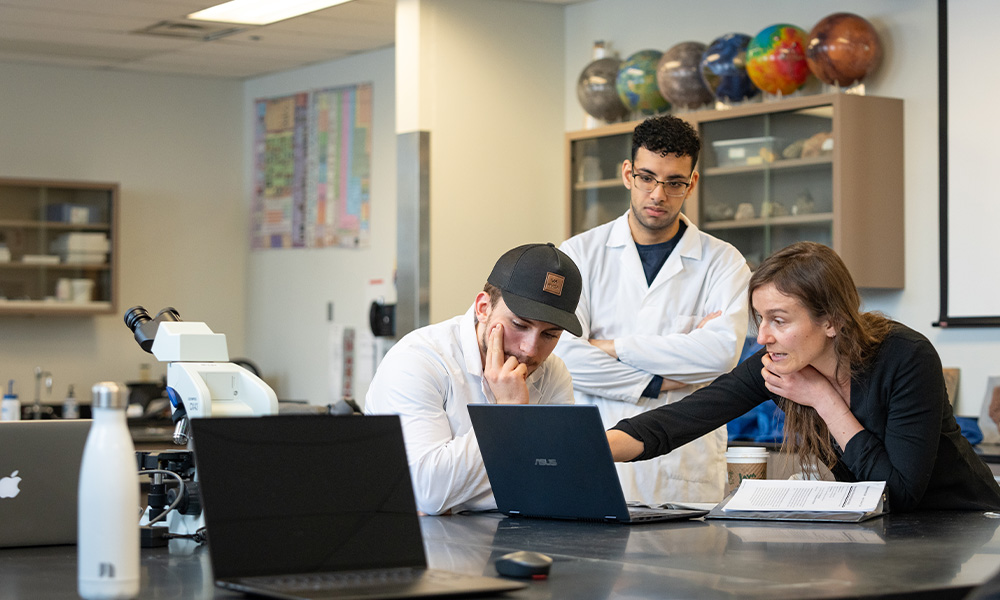 A woman points to something on a laptop screen while two male students in lab coats observe.