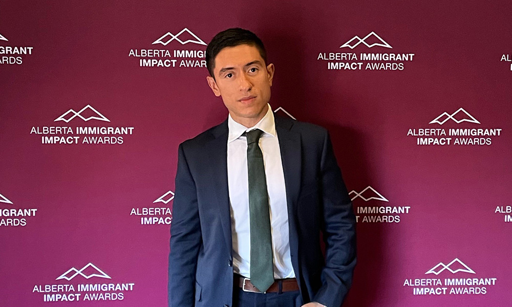 Carlos Flores stands against a maroon background with the logo of the Alberta Immigrant Impact Awards