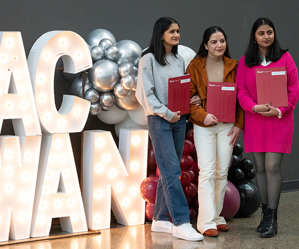 Three women stand in front of a display of balloons and large, lit-up words spelling MacEwan