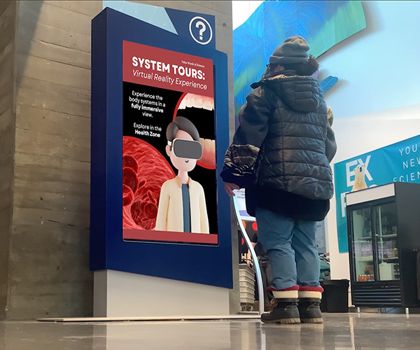 A person in a winter jacket stands in front of a display in the Telus World of Science that says System Tours