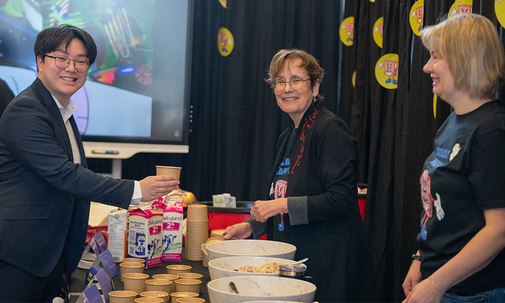 A woman smiling at the camera behind a table filled with cereal and milk, and a man also smiles at the camera while holding a paper bowl in his outstretched hand
